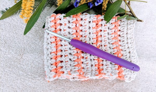 crochet washcloth in orange and off-white colour stripes, with purple crochet hook and flowers in the background