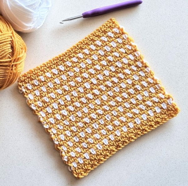 crochet blanket square in mustard and white with a crochet hook on white surface
