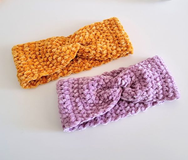 Two crocheted twist ear warmers in mustard yellow and soft lilac velvet yarn, laid flat on a white surface &mdash; a fast and cozy crochet beginner pattern perfect for fall and winter.