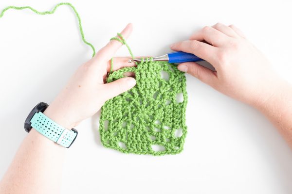 green crochet square on white background with hands and blue crochet hook