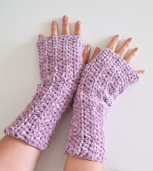 A pair of crocheted fingerless gloves in soft lilac velvet yarn, worn on both hands against a white background, showing a chunky textured stitch and a comfortable wrist-to-palm length fit.