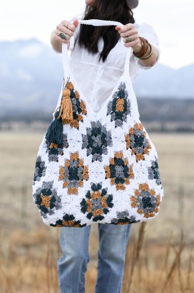 A woman in the background holding a crochet granny square bag in white, grey, brown, and dark blue colors.
