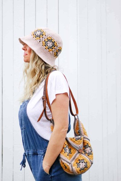 A woman in jeans overalls and a white t-shirt wearing a crochet granny square hat with a granny square crochet bag over her shoulder.