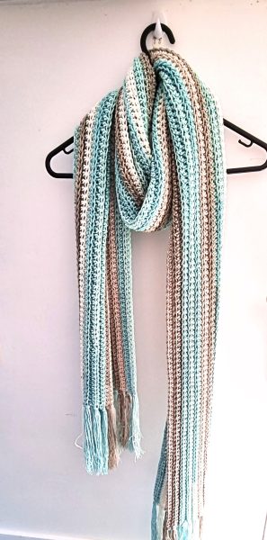 A long crocheted striped scarf in soft teal, cream, and tan with fringe ends, draped over a black hanger against a light wall, showing a delicate stitch texture.