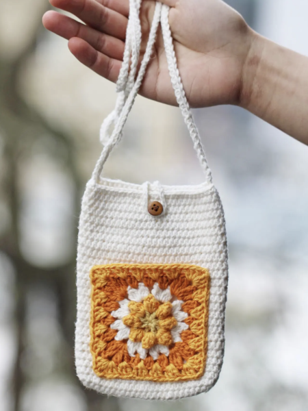 A hand holding a small white bag with a brown, yellow, and white granny square on the front.