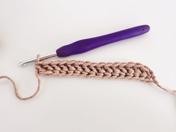a sample of foundation herringbone half double crochet row, showing 15 foundation stitches in light brown colour and a purple crochet hook. All on a white background.