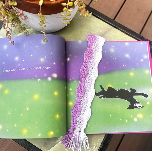 A crocheted bookmark in purple and white with twisted stripes and tassel ends, resting on an open illustrated book with a colorful green and purple night sky page, with a trailing plant and terracotta pot in the background &mdash; a sweet and quick crochet beginner pattern.