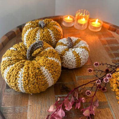 Three crochet pumpkins in yellow and white stripes on a wooden table with candles and dried flowers.