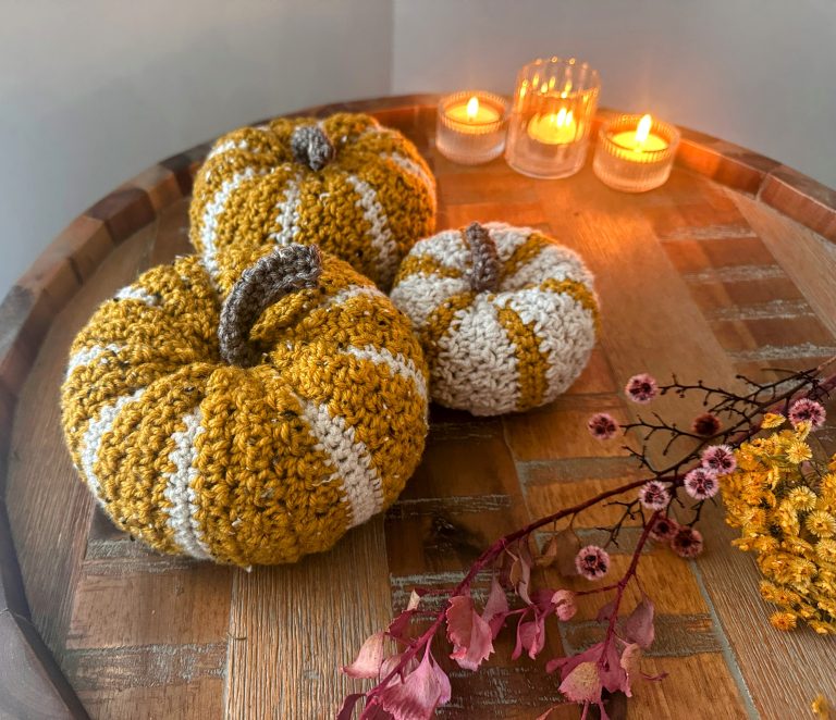 Three crochet pumpkins in yellow and white stripes on a wooden table with candles and dried flowers.