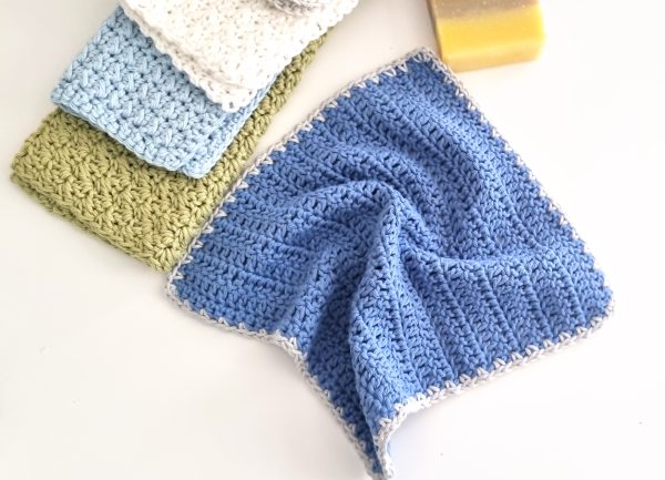 Four crocheted washcloths in blue, white, sage green, and gray, each with a contrasting border, arranged on a white surface next to a bar of soap &mdash; a practical and pretty crochet beginner pattern for the bathroom or kitchen.