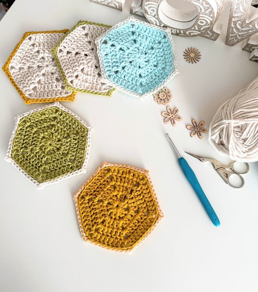 Five crocheted hexagon coasters in light blue, cream, mustard yellow, and sage green, each with a contrasting border, arranged on a white surface alongside a teal crochet hook, small wooden flower embellishments, and a ball of white yarn.