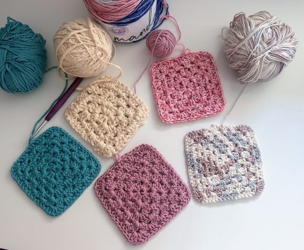 Five crocheted granny squares in teal, cream, pink, dusty rose, and variegated gray-blue &mdash; a great crochet beginner pattern &mdash; arranged on a white surface with matching yarn balls and a purple crochet hook.