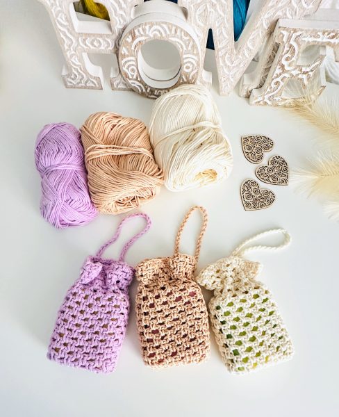 Three crocheted soap saver pouches in lavender, beige, and cream cotton yarn, shown with matching yarn skeins and decorative heart embellishments.