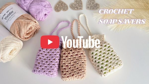 Three crocheted soap saver pouches in lavender, beige, and cream cotton yarn, shown with matching yarn skeins and decorative heart embellishments.